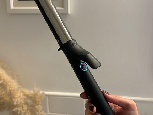 Hand holding the GHD Curve Hair Curling Iron in black with a small illuminated circular button under the extension that opens and closes the barrel