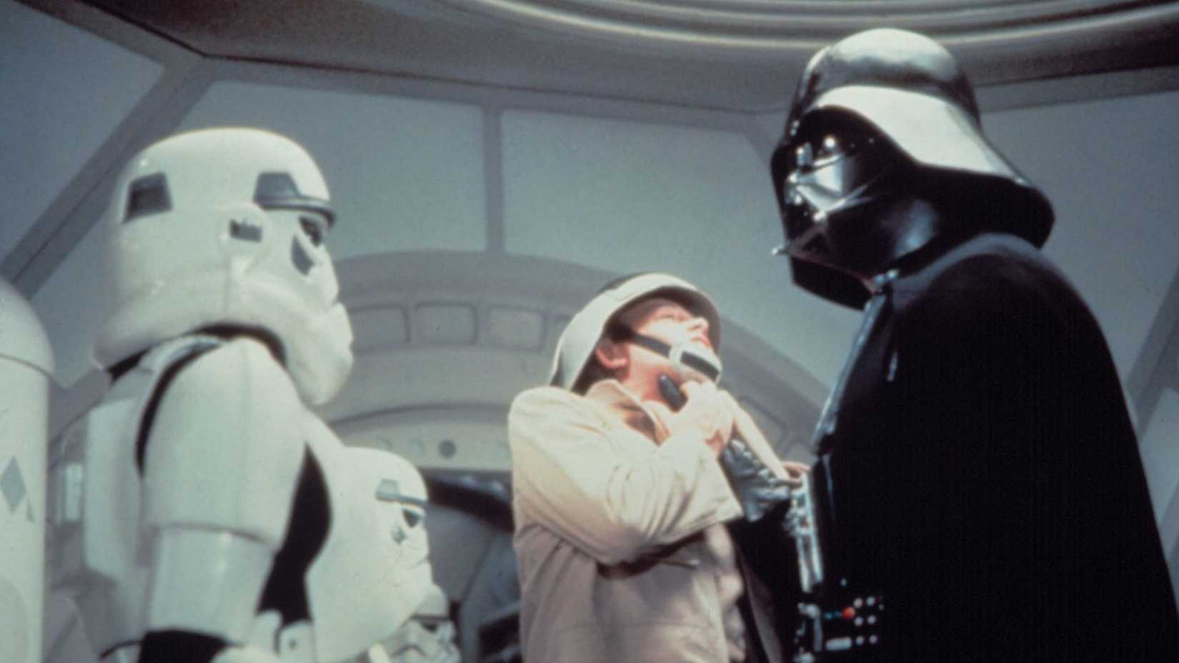 Darth Vader grasps a rebel by the throat and lifts him up. An Empire stormtrooper stands by, watching. From Star Wars: A New Hope.