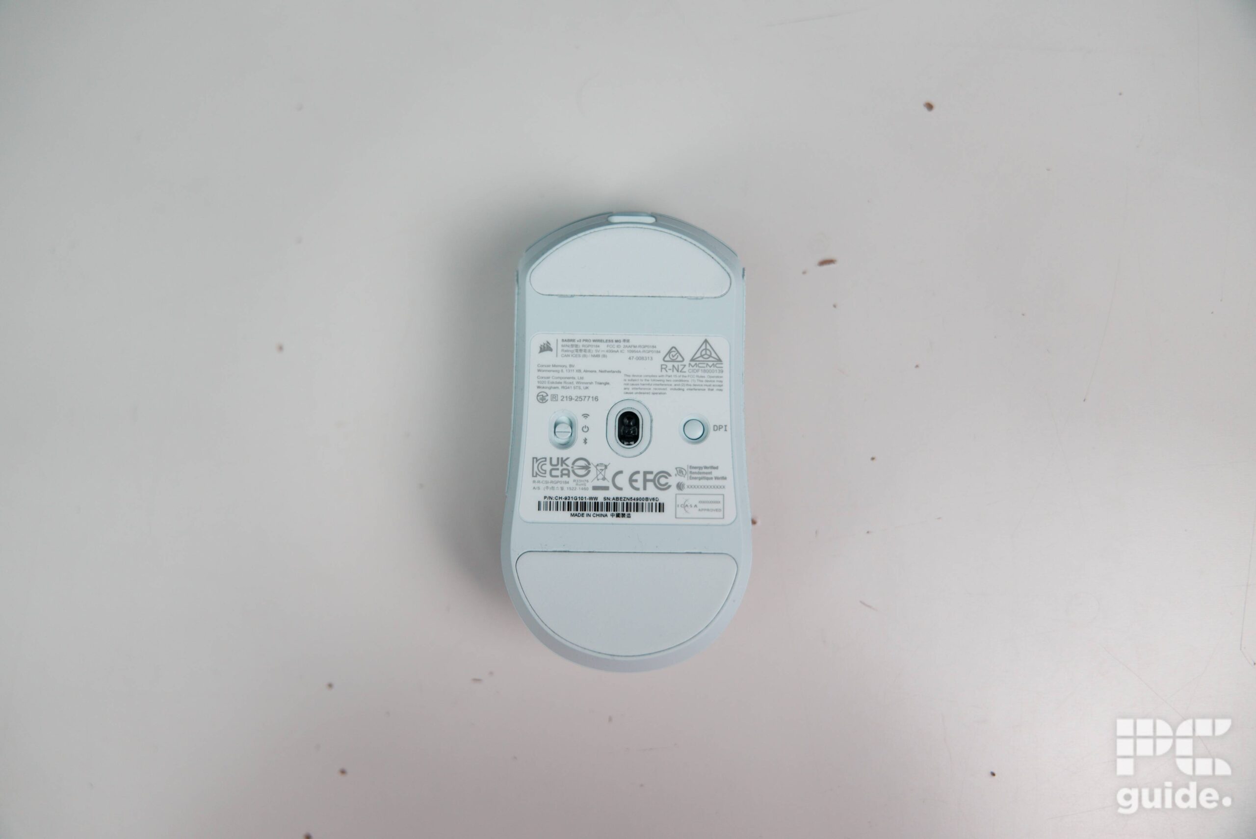 A top down shot of the underside of the white Corsair Sabre v2 PRO Wireless MG. Showing the skates, sensor, buttons, and sticker with specs.